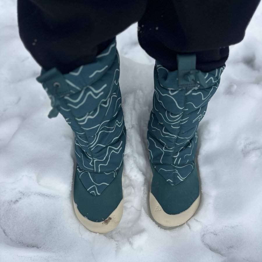 Top down look at a pair of warm winter waterproof kids snow boots. Vivobarefoot Tracker AT for kids in teal with a wide toe box and flexible outsole that moves with you. The boots are being worn on very snowy ground.