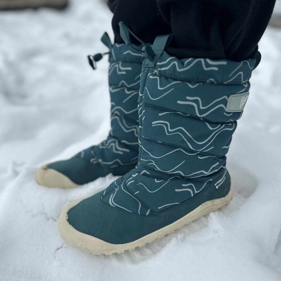 Side view showing a pair of zero drop wide toe box youth snow boots from Vivobarefoot. The Tracker AT is a warm waterproof boot being worn on very snowy ground.
