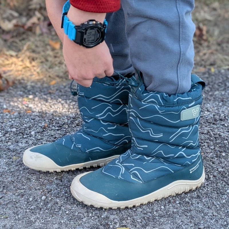 Side view of a pair of Vivobarefoot Tracker AT Snow Boots being worn by a young boy. The teal boots are warm and waterproof and have a cinchable top