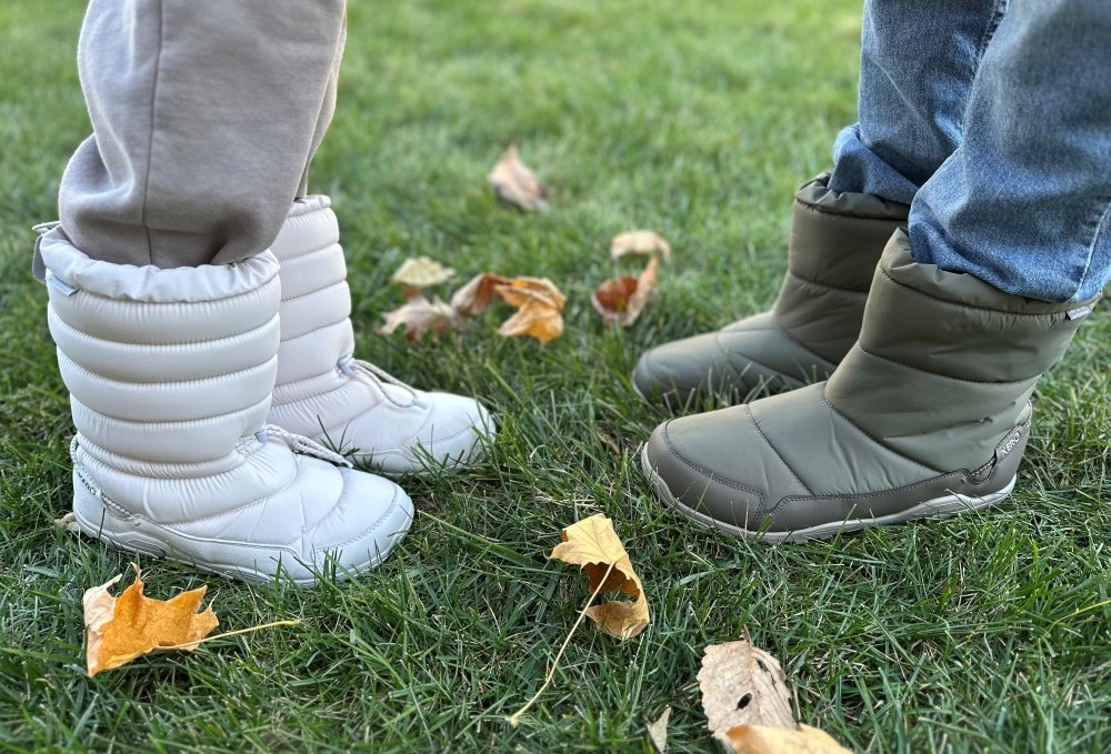 Two people standing toe to toe on grass with fallen leaves. They are both wearing Xero Alpine warm winter pull on barefoot boots with a wide toe box. The person on the left is wearing a white pair, while the person on the right is wearing an olive green pair.