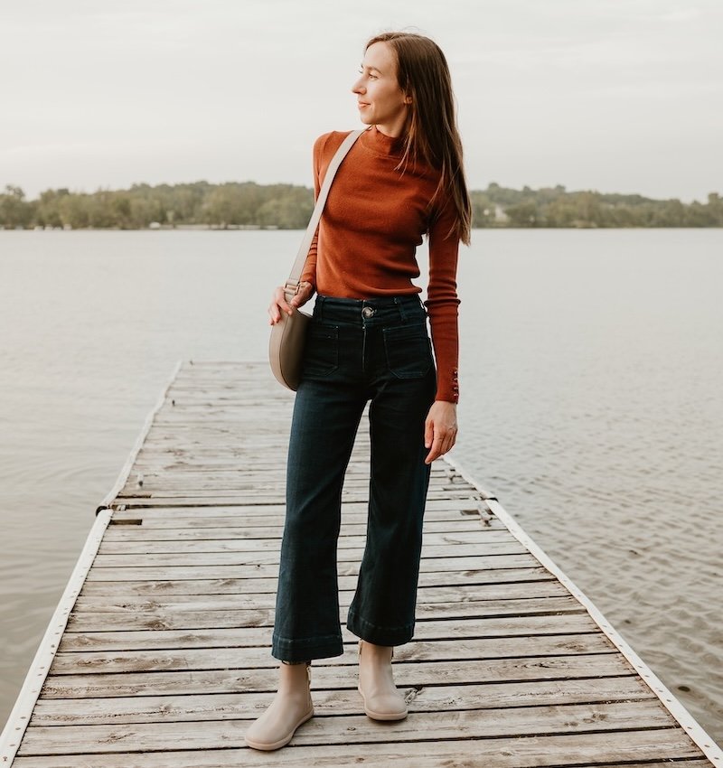 A woman in cropped dark denim and a burnt orange long sleeve top, standing on the dock of a lake with a handbag over her shoulder. She is looking off to the left with a slight smile on her face. On her feet are a pair of barefoot chelsea boots from Xero Shoes, a collabration with Anya's Reviews. The wide toe box taupe Tari II finishes the woman's outfit with a quiet elegance.