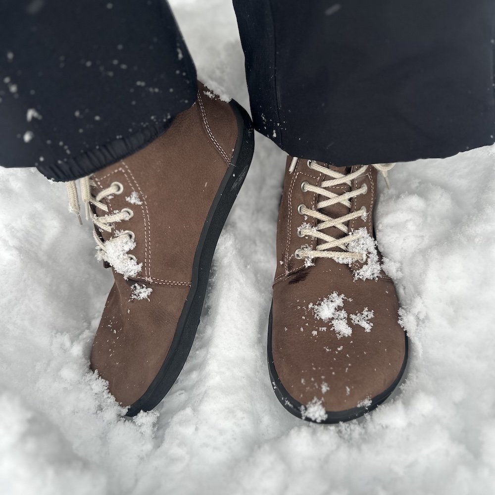 A front and side close up of a pair of Be Lenka Winter 3.0 barefoot wide toe box boots being worn in deep snow
