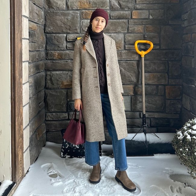 A woman standing on a snowy porch looking at the camera wearing a wool brown herringbone coat that falls below the knee, denim, and el naturalista wide toe box barefoot boots in light brown