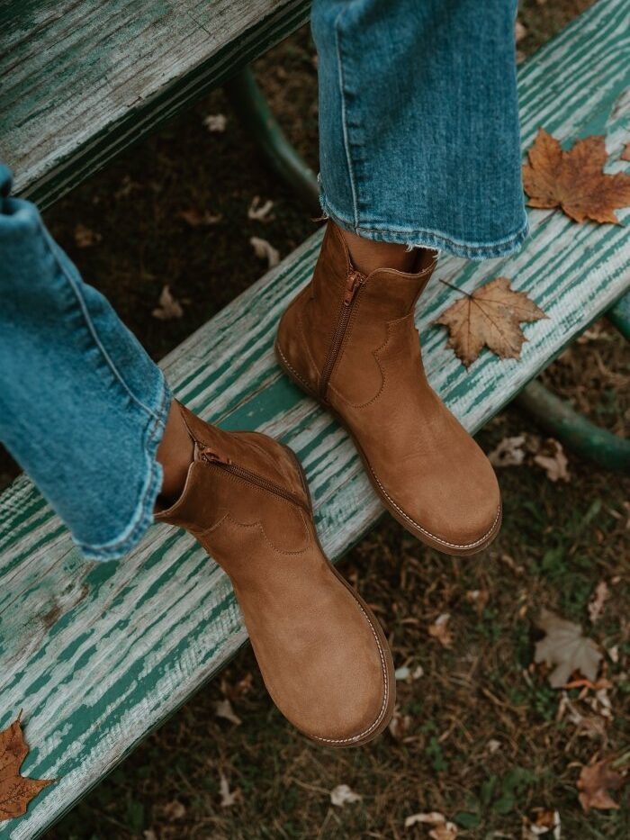 A close up top down view of a pair of Faye Boots by Anya in Cider colorway being worn by a person resting their feet on a worn picnic table bench. The wide toe box zip up western inspired boot is being worn with denim