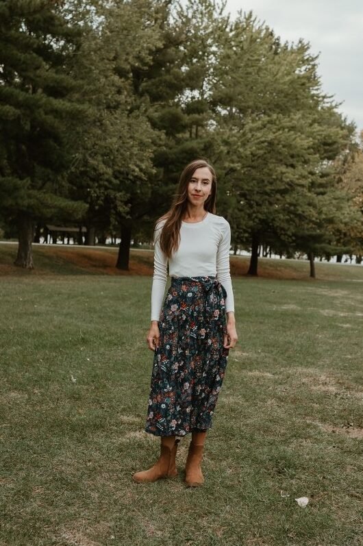 A full body view of a woman in a long navy floral skirt and long sleeve cream top. She is standing against a tree lined backdrop. She has finished her outfit with Faye Boots by Anya, a barefoot western inspired fashionable boot with a wide toe box.
