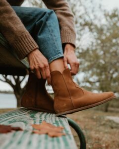 A side close up view of a pair of Cider Faye Boots by Anya with a zero drop outsole being put on by a woman sitting on a picnic table with her feet on the bench. The western inspired details are prominently displayed.