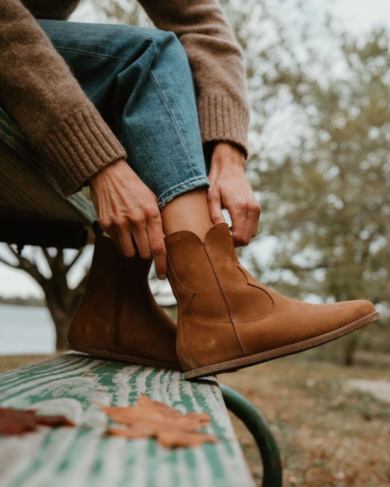 A side close up view of a pair of Cider Faye Boots by Anya with a zero drop outsole being put on by a woman sitting on a picnic table with her feet on the bench. The western inspired details are prominently displayed.