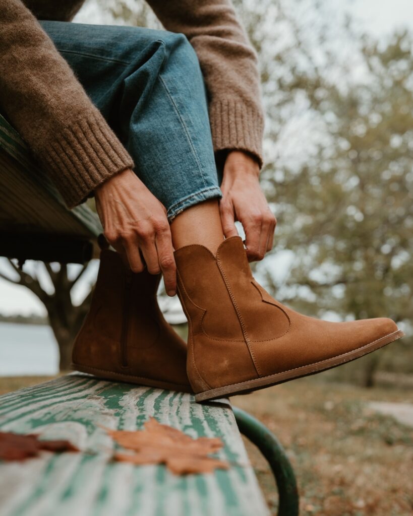 A side close up view of a pair of Cider Faye Boots by Anya with a zero drop outsole being put on by a woman sitting on a picnic table with her feet on the bench. The western inspired details are prominently displayed.