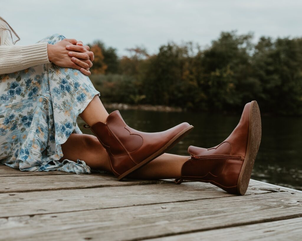 A knee down close up of a woman sitting on a wooden boat dock by a lake with her closer knee bent up and her feet resting on the wood. She has on a cream sweater and blue floral skirt paired with Faye Boots by Anya in smooth Cognac leather, a wide toe box western inspired zip up boots