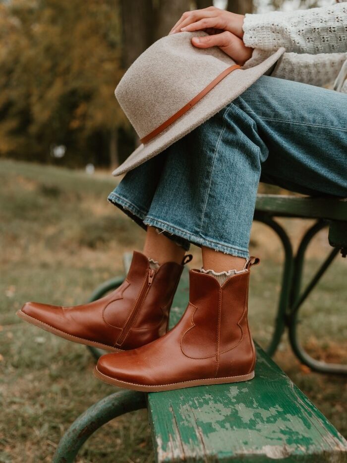Cropped view of a woman sitting on a picnic table with her feet resting on the bench. She has a wool hat resting on her knees, and her crochet knit sweater is just visible on her arms. Her feet are the focus on the photo, wearing a pair of Faye Boots in smooth Cognac leather, a wide toe box boot with western details