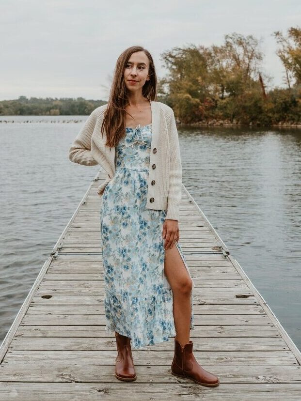 A full body view of a woman standing on the wooden dock of a lake wearing a blue floral dress and a cream cardigan. She has her left leg poking out of the dress slit and a pair of brown Cognac Faye Boots by Anya on her feet, a wide toe box dress boot.