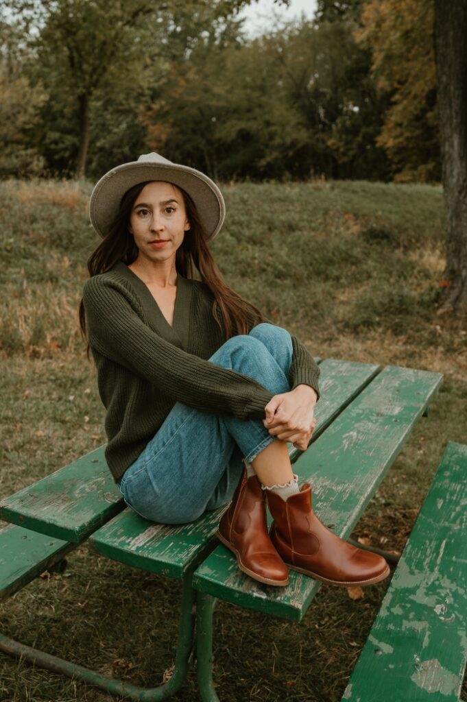 A full body view of a woman sitting on a picnic table with her feet crossed in front of her and her arms around her knees. She is wearing an olive green sweater and a wool oatmeal hat. She has on a pair of barefoot Faye Boots by Anya in Cognac brown, with western details, an easy on zipper, and a comfortable wide toe box.