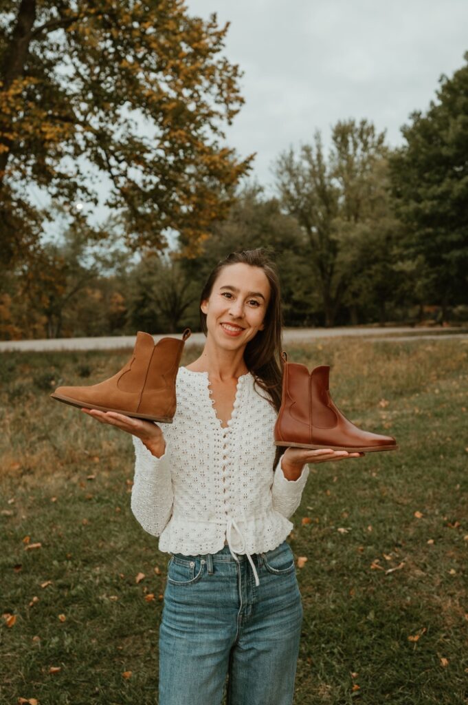 A cropped shot of a woman wearing a white crochet peplum sweater and jeans holding one Cognac and one Cider Faye boot by Anya in each hand.