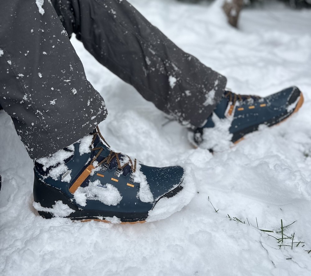 Close up side view of a pair of Lems Breck insulated wide toe box winter boots being worn by a man in deep snow.