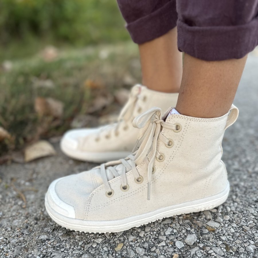 Side close up view of a pair of barefoot Tolos 2.0 wide toe box canvas high tops with a wide toe box and flexible outsole being worn on a pebbled path. A pair of plum colored linen pants are visible at the top of the frame.