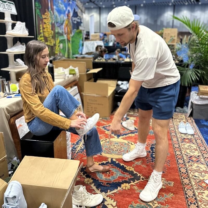 A booth at a barefoot shoe expo, where the owner of Tolos is helping a woman test out a pair of Tolos Transition shoes.
