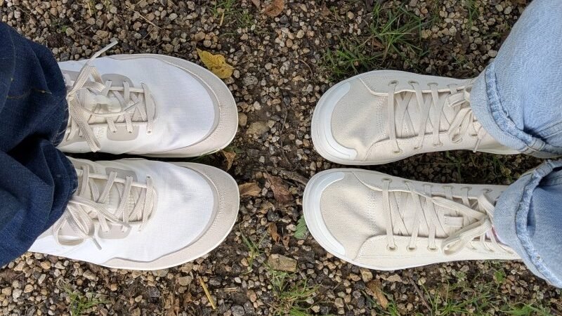 Two people's feet close up in Tolos wide toe box Transition models standing toe to toe on a grassy and gravel path.