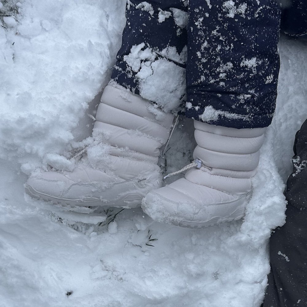 A pair of white Xero Alpine pull on puffer winter boots being worn by a person against a very snowy ground.