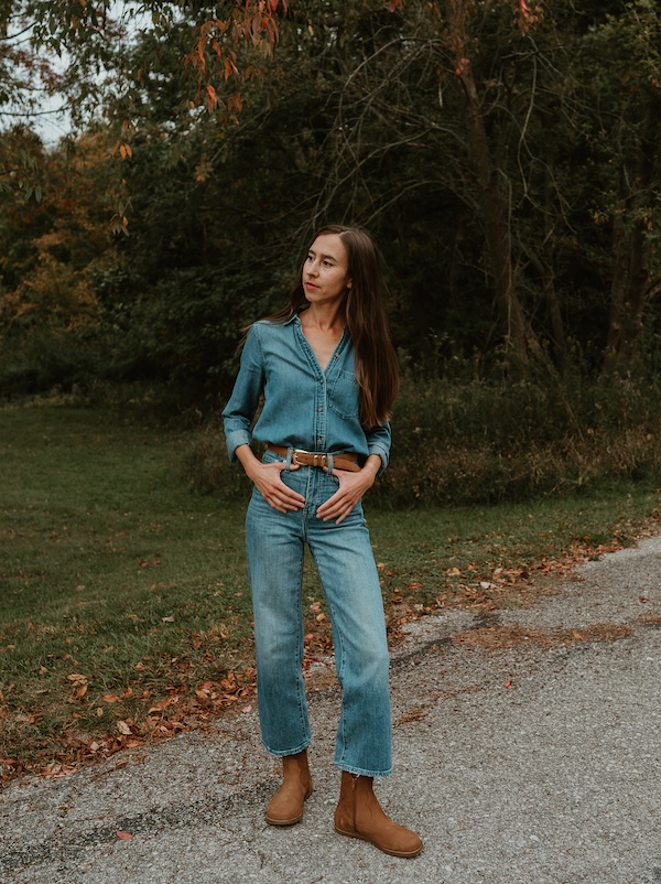 A woman standing in front of a grassy patch with fallen leaves wearing denim on denim with a tan suede belt and Cider nubuck Faye by Anya wide toe box barefoot boots