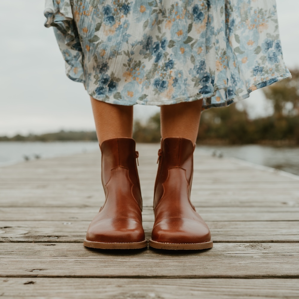 A close up front view of a person's calves and feet standing on a boardwalk with the bottom hem of a floral skirt visible. The person is wearing Faye by Anya foot shaped barefoot boots in cognac leather.