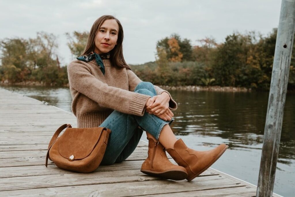 A woman sitting on the dock of a lake wearing a light brown sweater, jeans, and a blue silk scarf tied at her neck. She is holding her knees and her feet are crossed with a pair of Faye Boots by Anya in Cider resting against the wood boards. A matching leather handbag is resting against her hip.