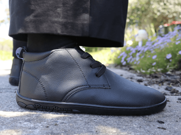 Side close up of a pair of black leather chukka boots for a barefoot school shoe option from PaperKrane. The person is standing outside on a sunny day with purple flowers in the background. The shoes are shown from the side with a good view of the flat flexible outsole.