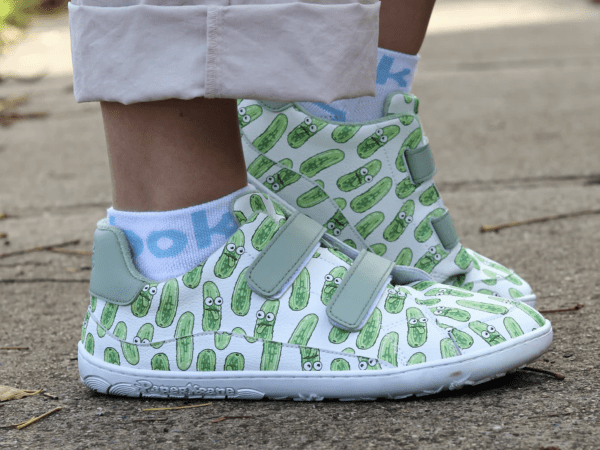 A close up side view of a pair of white leather Pickle print PaperKrane velcro barefoot sneakers. A young person is wearing them outside and the back foot is flexed up.