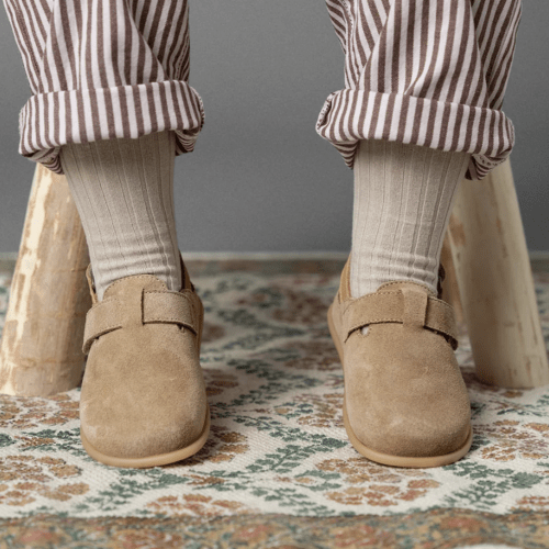 Front view of a small child's feet in a pair of Barebound suede clogs in tan being worn while sitting on a wooden stool.