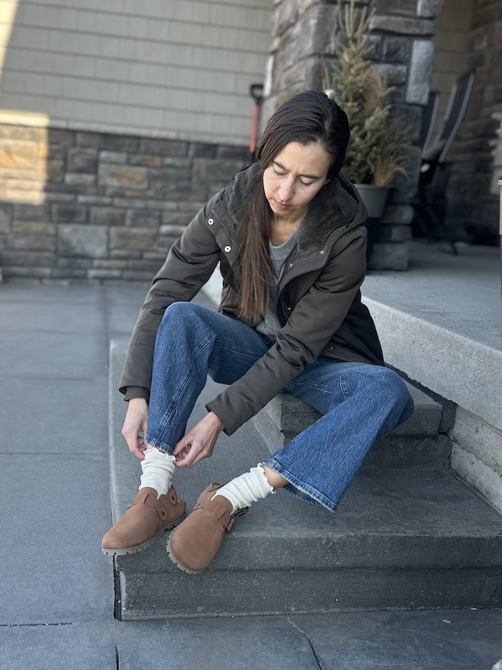 A woman sitting on her front steps putting on a pair of Barebound Ikaros tan Terracotta clogs with a barefoot outsole