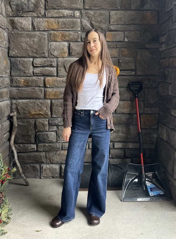A full body view of a woman wearing dark wide leg jeans, Barebound Ikaros barefoot clogs, and a brown sweater. She is standing in an alcove with her hand in her pocket.