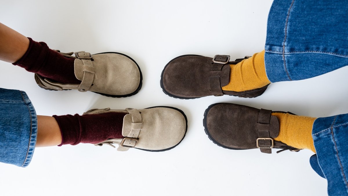 Top down wide image of two people's feet in Barebound Ikaros Brown clogs in brown and tan suede. Both are being worn with slouchy socks and wide leg denim