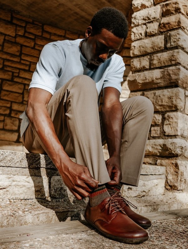 A man sitting on a curb wearing dress slacks, bending down to tug at him pant hem. He is wearing brown leather Groundies barefoot dress shoes for men.