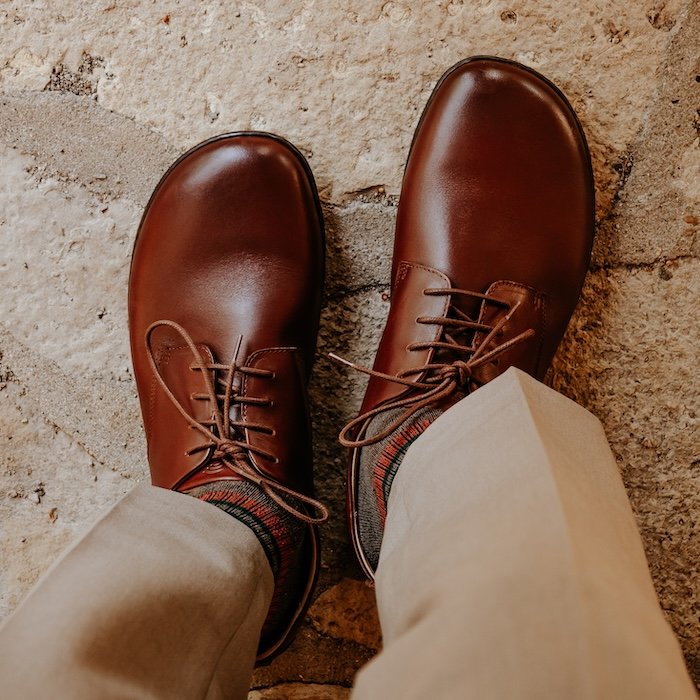 A top down view of a pair of feet standing on tan pavement wearing tan slacks and brown leather Groundies men's barefoot dress shoes