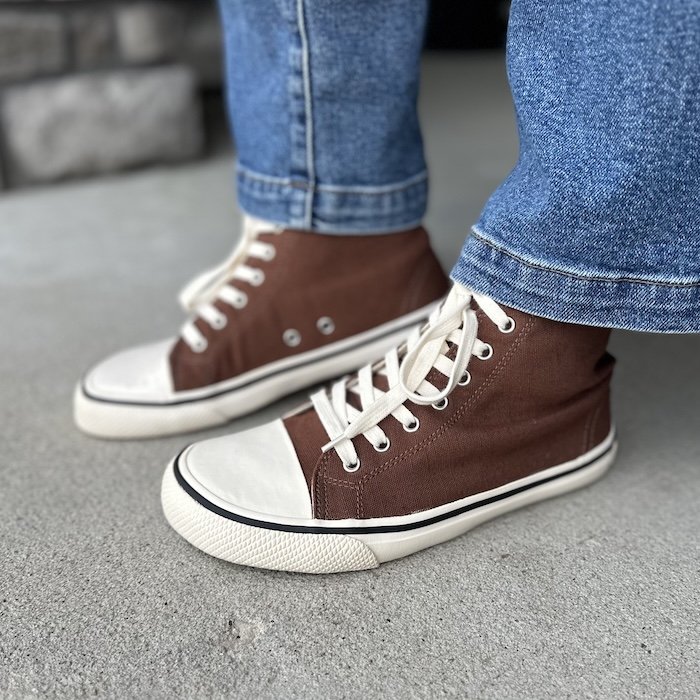 A close up left side view of a person wearing jeans with the Bohempia Orik brown and tan high top sneakers. The sneakers are a brown canvas high top with white laces and a light tan rubber toe and sole.