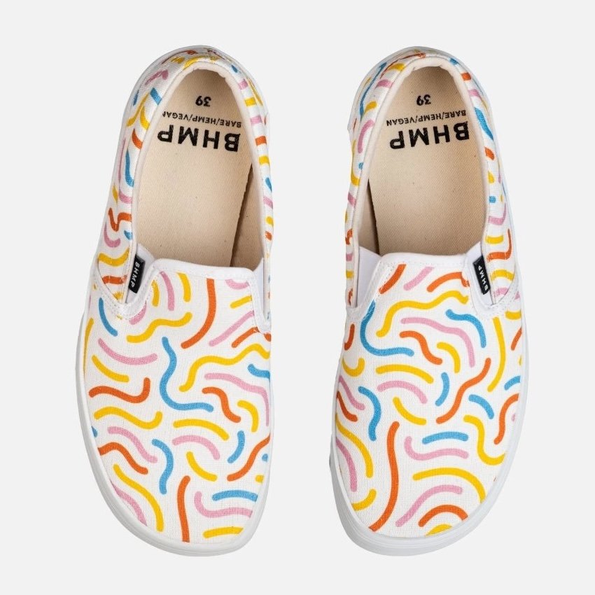 A top down view of the Bohempia Velik slip on sneakers in their Candy print on a white background. They are a white hemp canvas slip on sneaker with blue, yellow, orange, and pink squiggles all over.