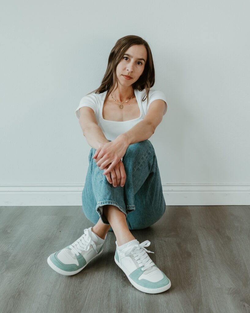 A person sitting on the floor with her legs crossed and arms resting on the knees. She has a slight smile on her face and is wearing a pair of Shapen ReWind barefoot sneakers in Jade and white leather.