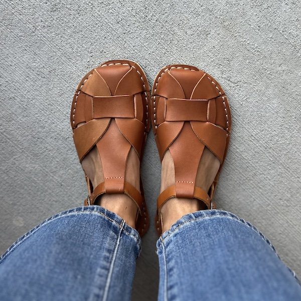 A close up top down view of a woman wearing jeans and the Crupon Abbie leather sandals in the Ambra color. The Abbie sandals are a brown fisherman style sandal with a woven design over the top of the foot.