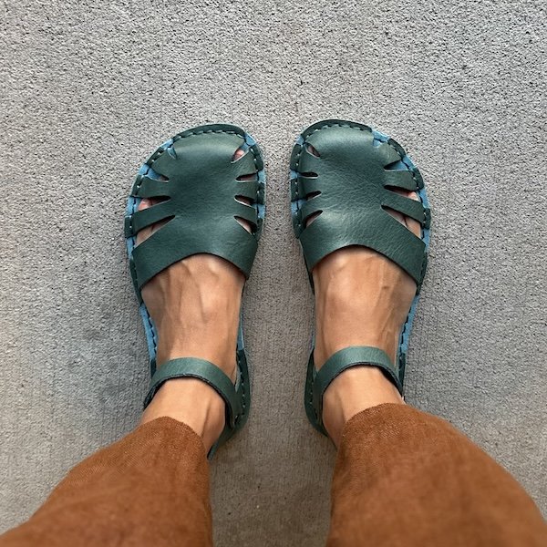 A close up top down view of a person wearing brown linen pants and the Crupon Monstera leather sandals in their Avion color. The sandals are a jade green color with a turquoise footbed, and have an ankle strap and cutouts over the toe area.