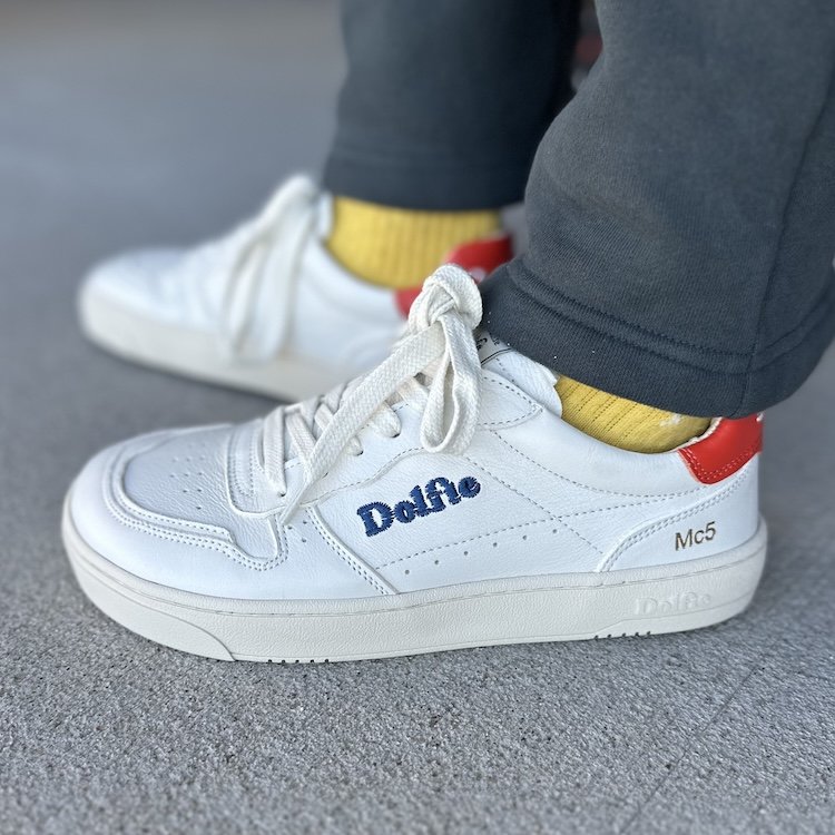 Side view of a pair of Dolfie Paradise white Mc5 sneakers with a wide toe box and zero drop cushioned sole. The embroidered Dolfie logo is on the side in blue, and the heel of the sneaker is red.