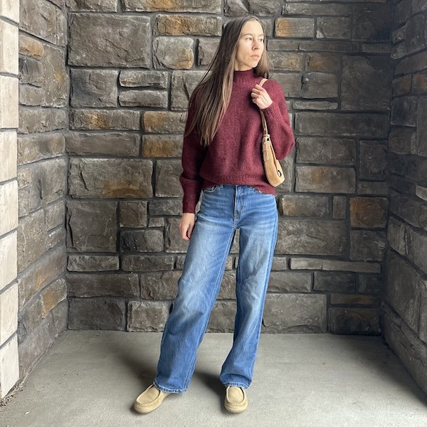 A full body view of a woman looking off camera in a pair of mid wash denim, a burgundy sweater, and Groundies Cricket barefoot shoes.