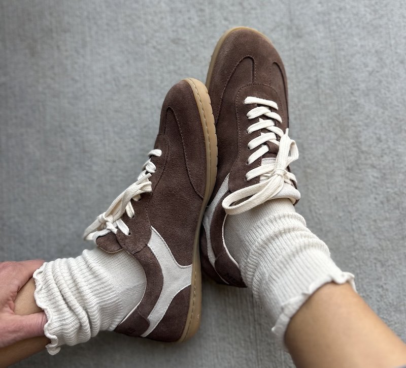 A close up view of a person wearing the Mukishoes Lyra Rheia sneakers with white ruffled crew socks. The sneakers are brown suede and have a white detail below the ankle.