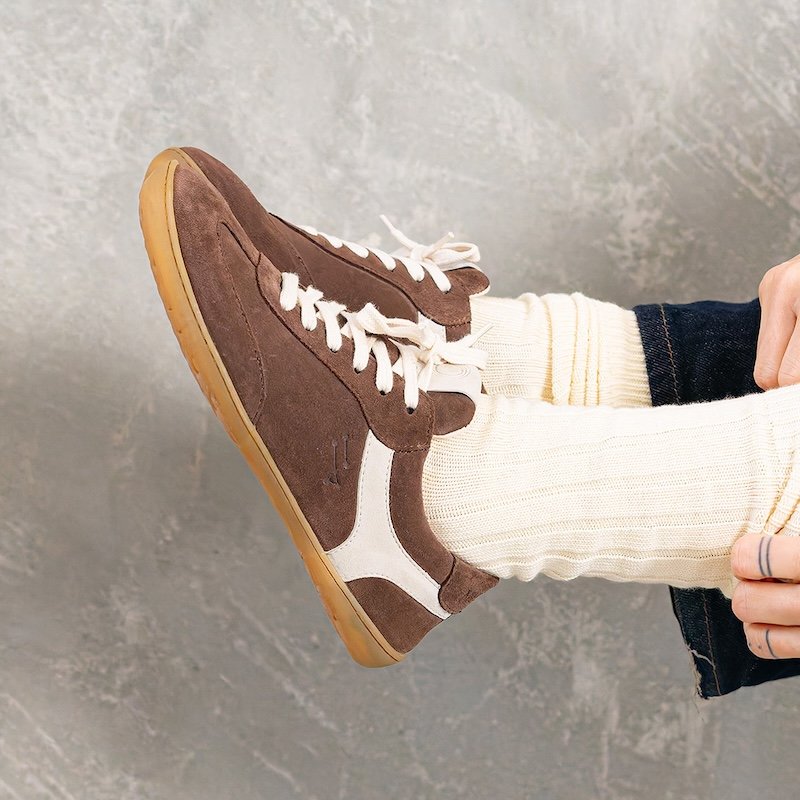 A side view of a person's feet in Mukishoes Lyra Rheia brown suede barefoot sneakers with soft flexible outsole. The shoes are suspended in the air, and a person's hands are pulling up the cream colored socks being worn.