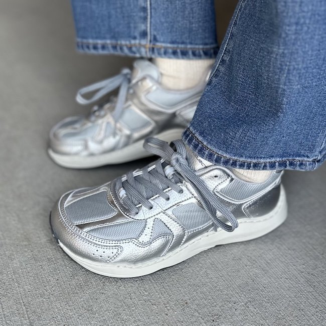 A close up left side view of a person wearing jeans and the Ohne Project Retro Run silver sneaker. The sneakers are a silver modern running sneaker with reflective details and mesh panels.