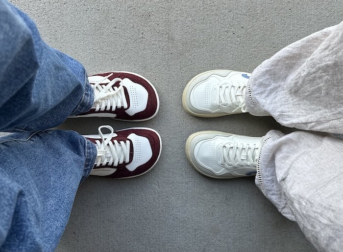 A top down view of two people's feet wearing the Ohne Project barefoot sneakers. The person on the left is wearing blue jeans with the red wine color sneakers, which are a white leather with burgundy details. The person on the right is wearing white linen pants with the heritage jeans color sneaker, which is a white leather sneaker with light grey and light blue details.