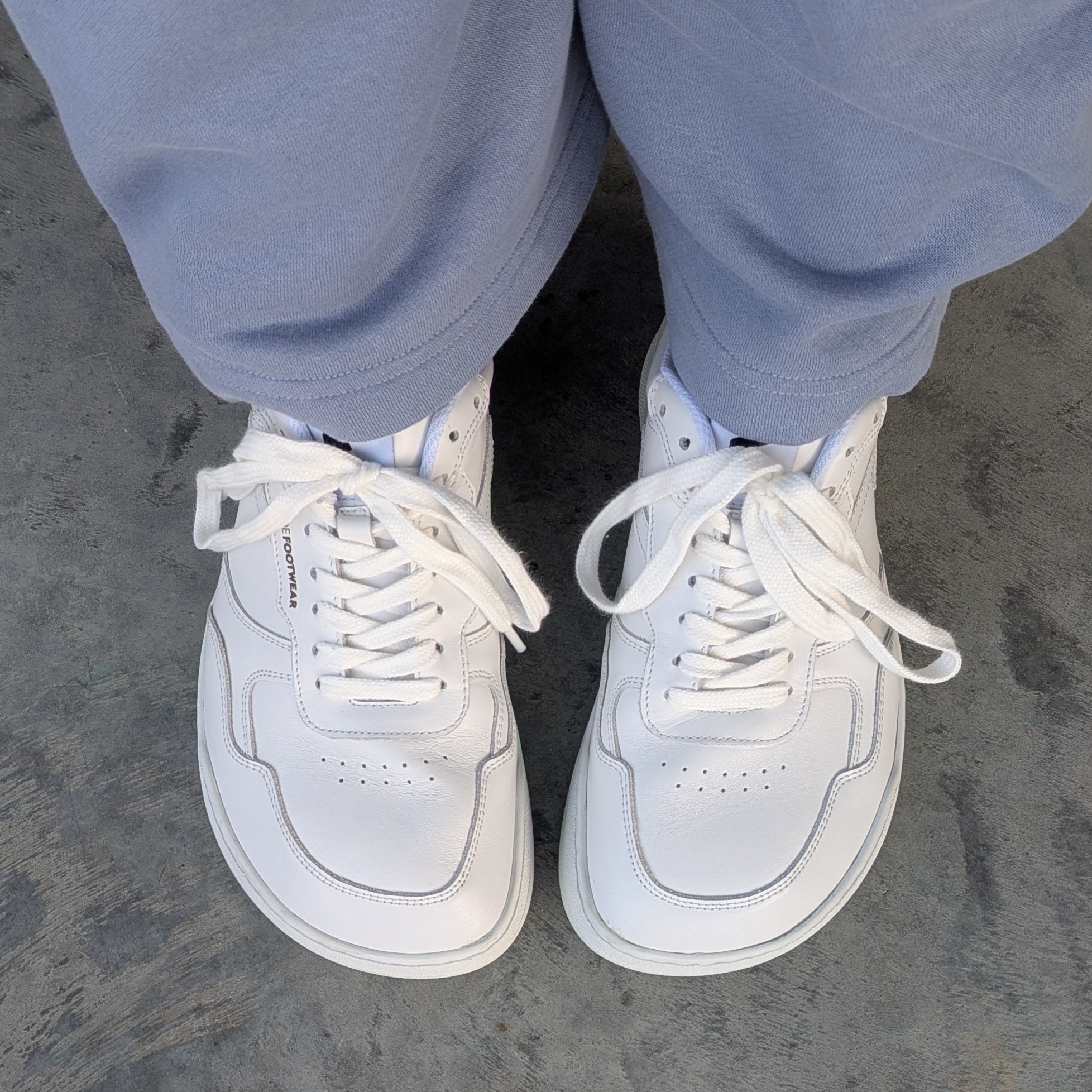 Top down view of a person's feet in extra wide toe box sneakers from Wyde Footwear. The Nova in all white leather is shown with lavender casual pants.