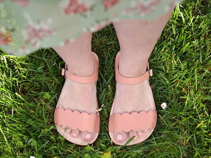 A top down view of a person standing in grass while wearing a green floral dress and the Softstar Dahlia leather sandals in wide width. The sandals are a light brown leather, with a buckled ankle strap and a panel over the toes that has a scalloped design.