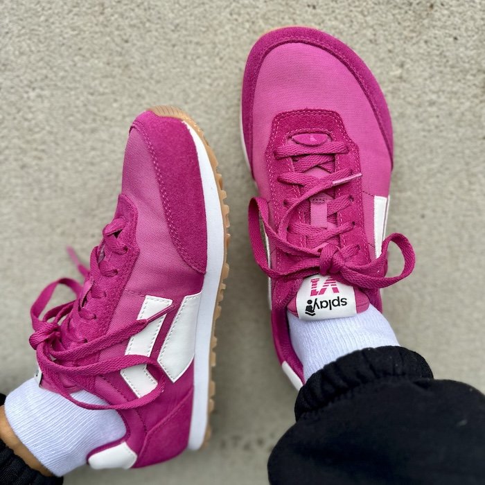 A close up of a pair of feet resting on concrete wearing bright pink retro Splay Shoes Runner barefoot sneakers