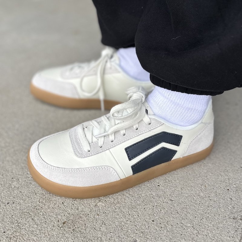 A close up side view of a pair of feet standing on concrete wearing Splay Shoes 101 barefoot sneakers in white leather with blue accents