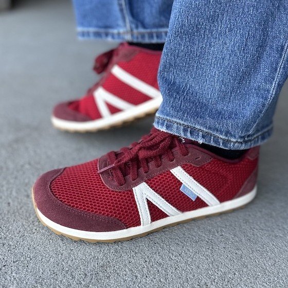 Side close up view of a woman's feet in Xero Wynn retro red barefoot sneakers with white accents. The zero drop outsole is at the forefront of the image, and a pair of midtone denim rests at the ankle.