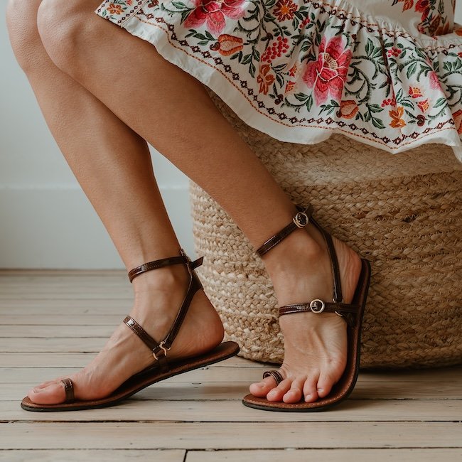 A close up of a pair of High Feels patent strappy barefoot sandals in dark brown. The person is sitting on a woven jute cushion and wearing a pretty floral dress that's just visible at the top of the frame. The left foot is flexed up to show outsole flexibility.